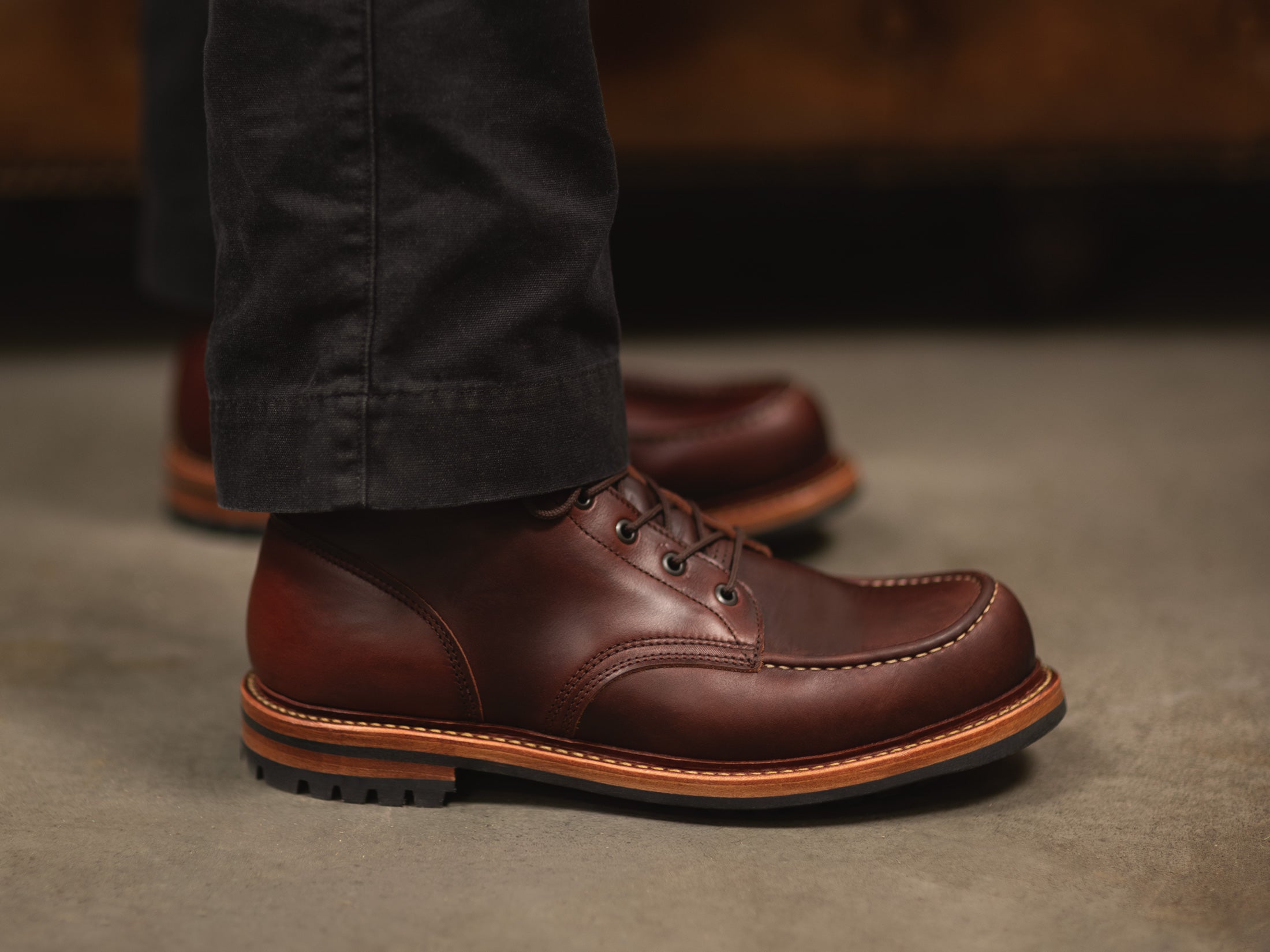 side view of rich brown moc toe boots standing near leather couch